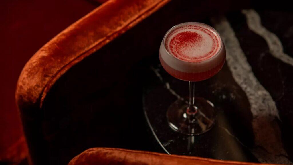 A cocktail with a foamy top and red powder is served in a coupe glass on a dark marble table, surrounded by orange velvet seating—the perfect setting for an intimate Valentine’s Day celebration or festive Lunar New Year toast.