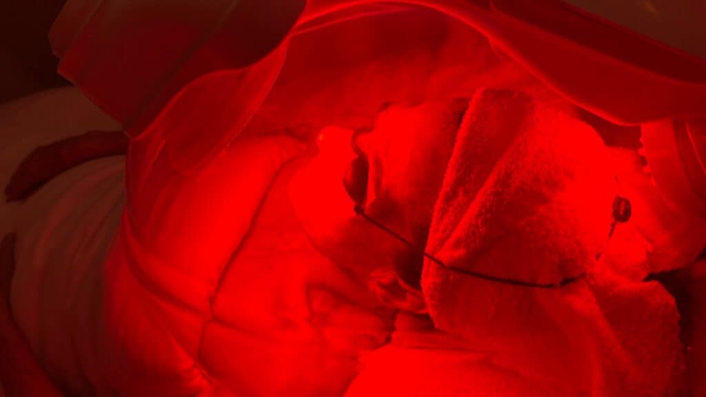 A person lies under a red LED light therapy device, with their head wrapped in a towel and eyes protected by goggles.