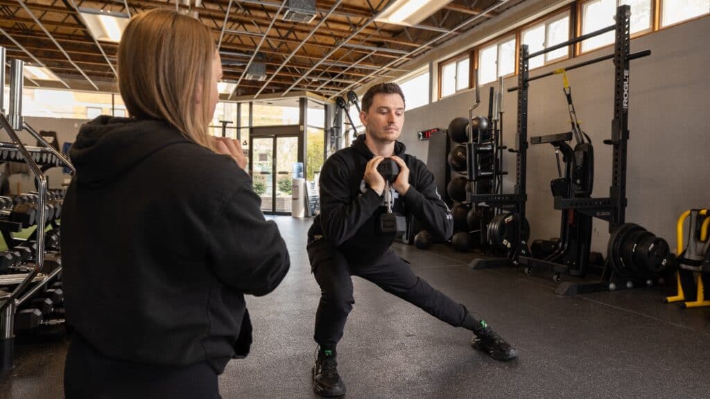 A man performs a side lunge with a kettlebell in a gym while a trainer observes him.