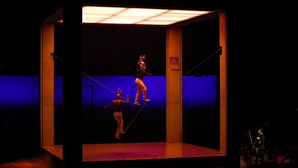 Two performers balance on tightropes inside a large illuminated cube structure at ECHO, with an audience watching in the background.