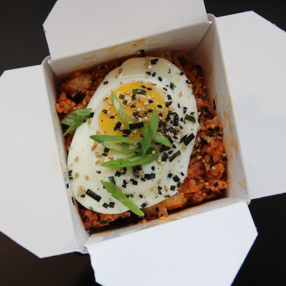 A takeout box filled with fried rice topped with a sunny-side-up egg, sliced green onions, sesame seeds, and seaweed flakes.