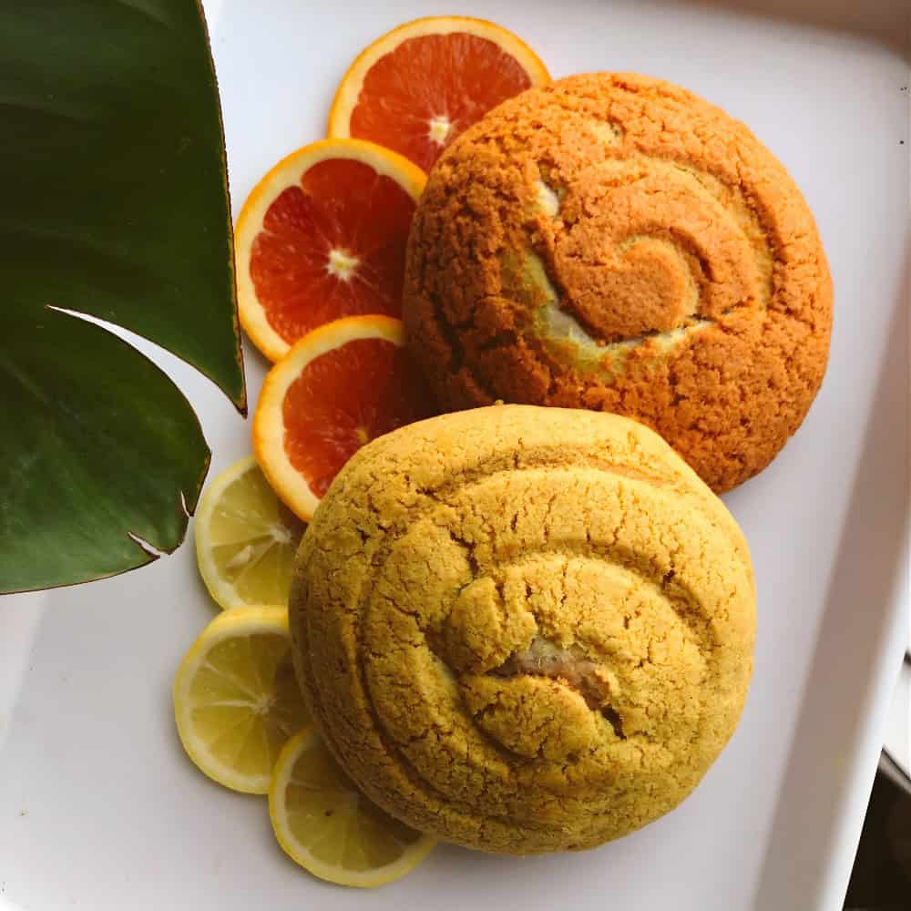 Two concha pastries on a white tray with slices of orange and lemon, partially covered by a large green leaf in the corner.