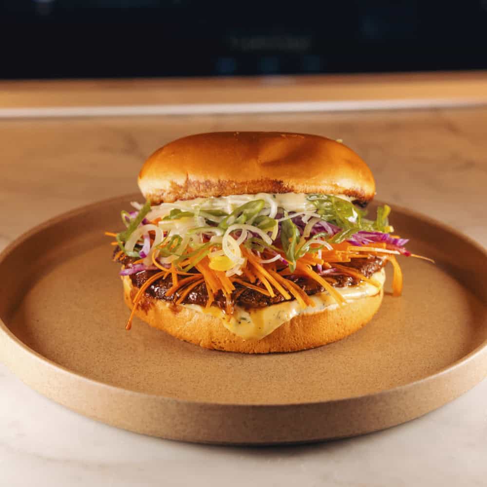A sandwich with shredded meat, sliced carrots, lettuce, and sauce in a brioche bun, served on a round, beige plate on a marble surface.