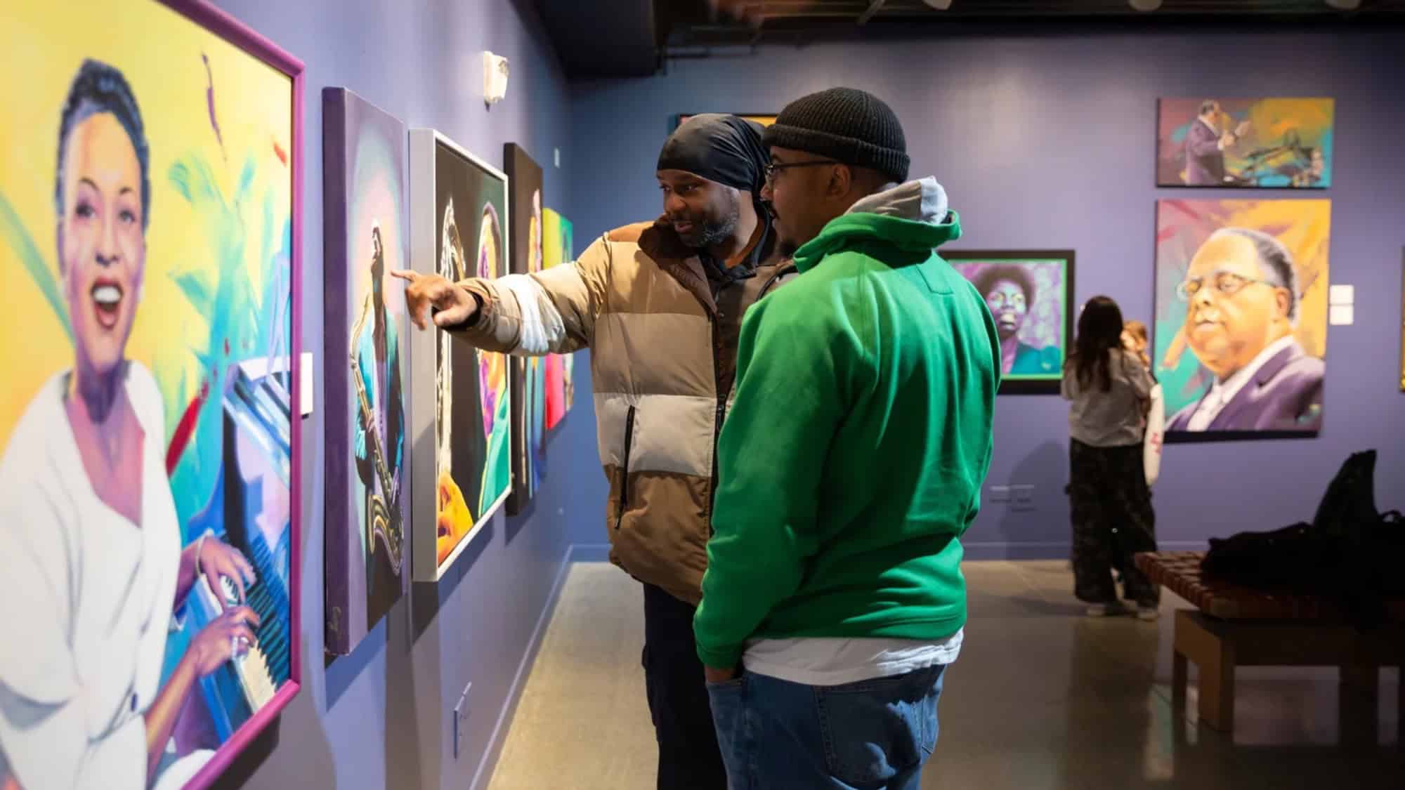 Two people observe and discuss colorful portrait paintings in an art gallery, while others view artworks in the background.