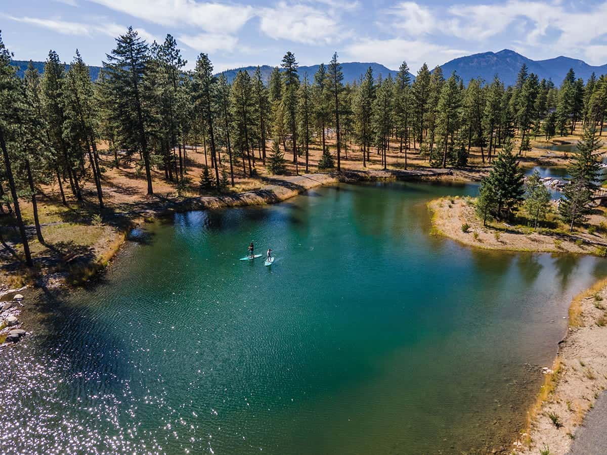 Two people paddleboard on a small, clear lake surrounded by pine trees at Suncadia, with mountains in the background—an idyllic scene to build your home in your style and enjoy life immersed in nature.