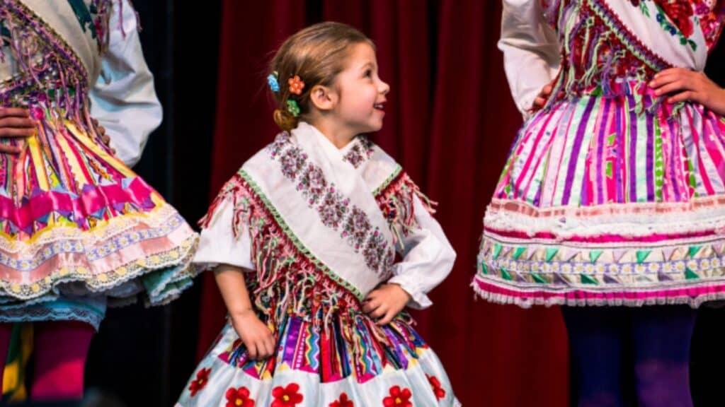 A young girl in a colorful, traditional folk costume stands with hands on her hips, smiling and looking at an adult beside her, against a red curtain backdrop.