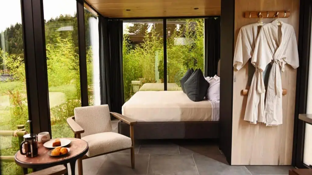 Modern bedroom with floor-to-ceiling windows, a bed with white linens and gray pillows, a chair and table with breakfast, and two robes hanging on the wall. Lush greenery is visible outside.