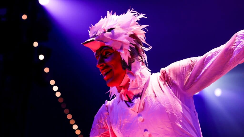 Performer in a white bird costume with feathers and beak mask poses under purple stage lights.