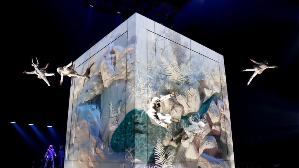 Four acrobats in white costumes perform mid-air stunts around a large illuminated cube with nature-themed projections on its surface, under stage lighting.