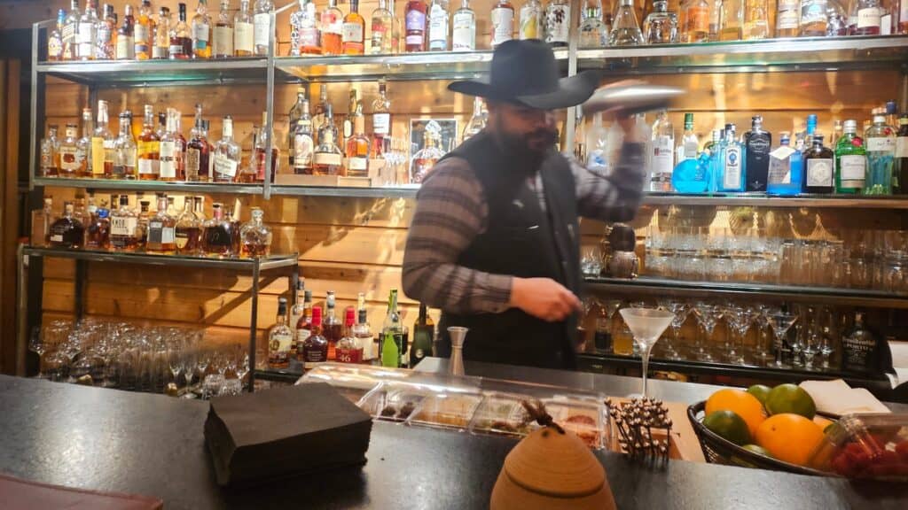 A bartender in a cowboy hat shakes a cocktail behind a bar stocked with bottles and glasses, with fruit and napkins on the counter.