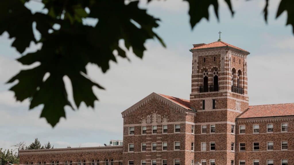 A brick building with a prominent bell tower and red-tiled roof, partially framed by tree leaves in the foreground under a cloudy sky—a perfect spot for romantic Valentine’s Day getaways.