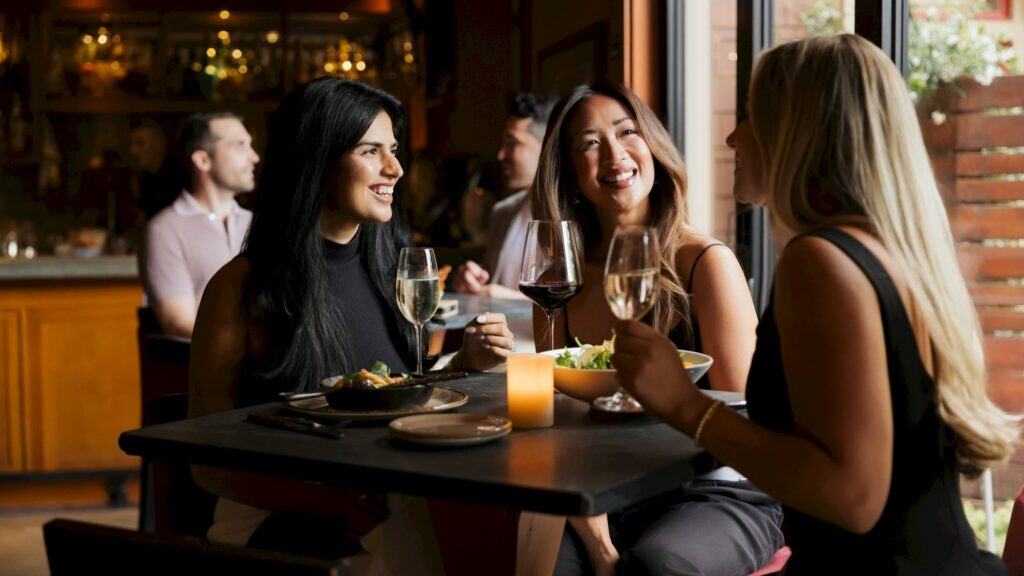 Three women sit at a restaurant table by a window, enjoying wine and lively conversation on Valentine’s Day, while two people relax at another table in the background.
