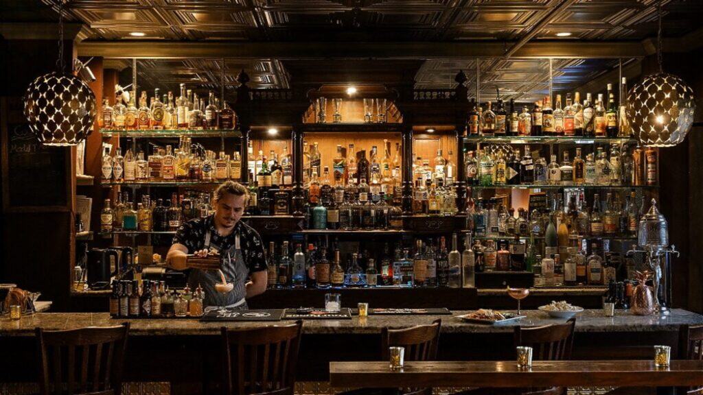 A bartender prepares a drink behind a well-stocked bar with shelves of liquor bottles, dim lighting, and empty chairs—perfect for cozy Valentine’s Day getaways or celebrating Lunar New Year in style.