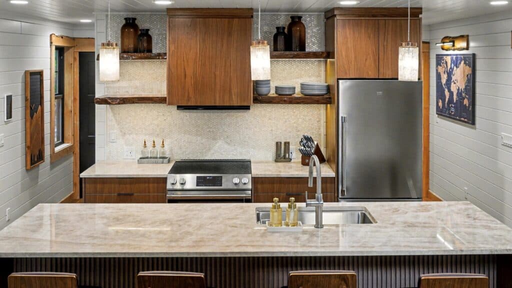 Modern kitchen with wood cabinets, stainless steel appliances, marble countertops, open shelves with dishes, and decorative bottles on display above the stove—beautifully built into the trees for a serene, nature-inspired setting.