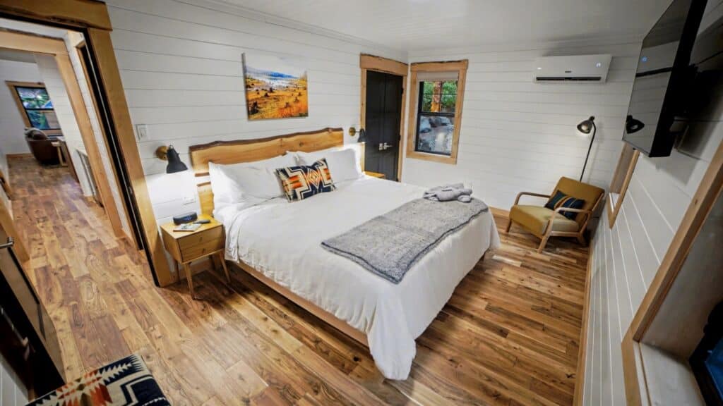 A modern bedroom with a wooden floor, a bed with white linens, two bedside tables, a chair, wall-mounted lamps, a painting, and an air conditioner.