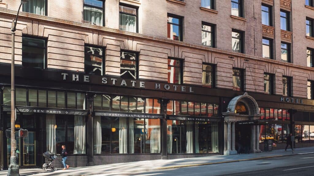 The exterior of The State Hotel, featuring large windows, the hotel sign, and neighboring Ben Paris restaurant on a city street—perfect for memorable getaways or celebrating special occasions like Valentine’s Day.