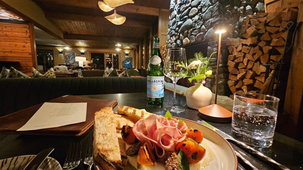 A plate of bread, vegetables, and cured meat sits on a table with sparkling water, a glass, a menu, and a flower vase; the background shows a cozy, wood-paneled lounge with a stone fireplace.