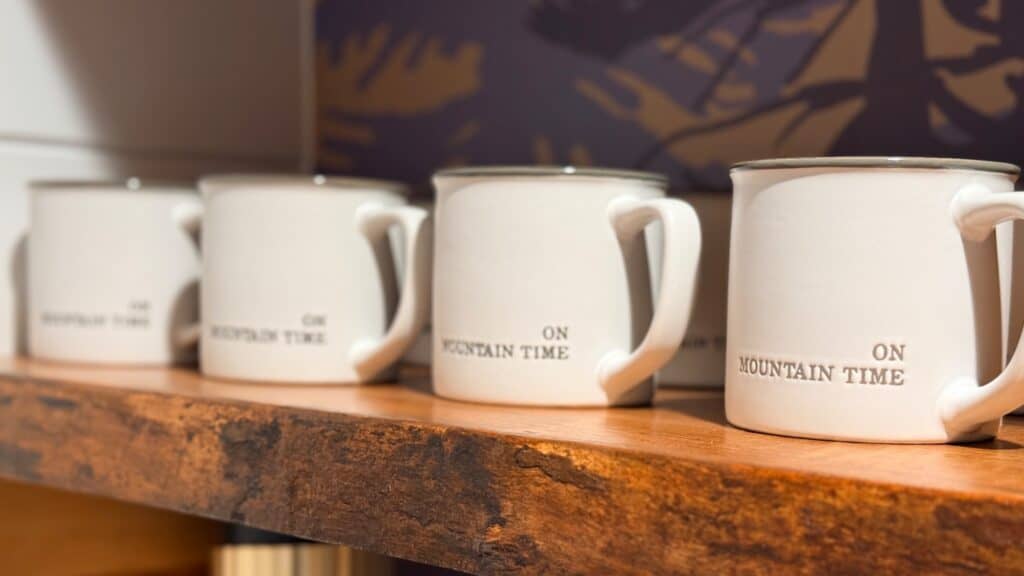 Four white ceramic mugs with the words "ON MOUNTAIN TIME" are lined up on a wooden shelf, perfect for cabins built into the trees.