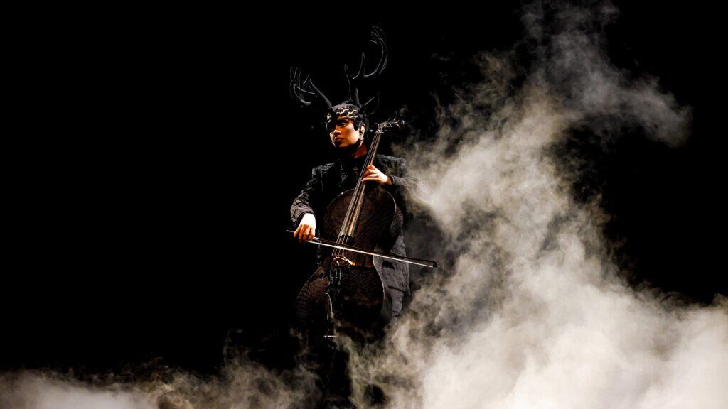 A cellist in dark clothing and an antlered headpiece plays a cello amid dramatic stage smoke against a black background.