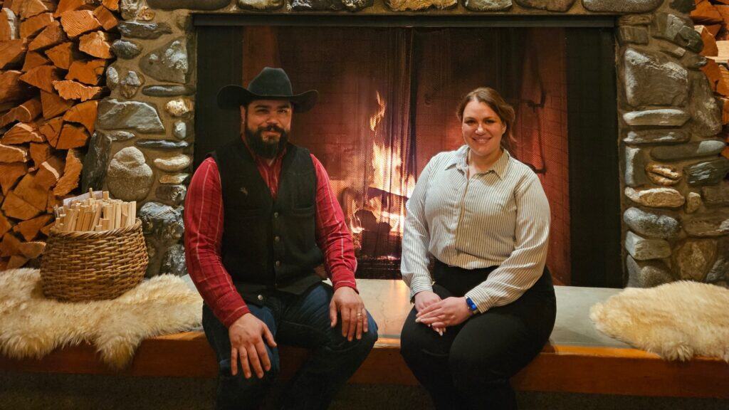Two people sit on a stone fireplace hearth with a fire burning behind them. A basket of wood and stacked logs are at the left, and both are smiling at the camera.