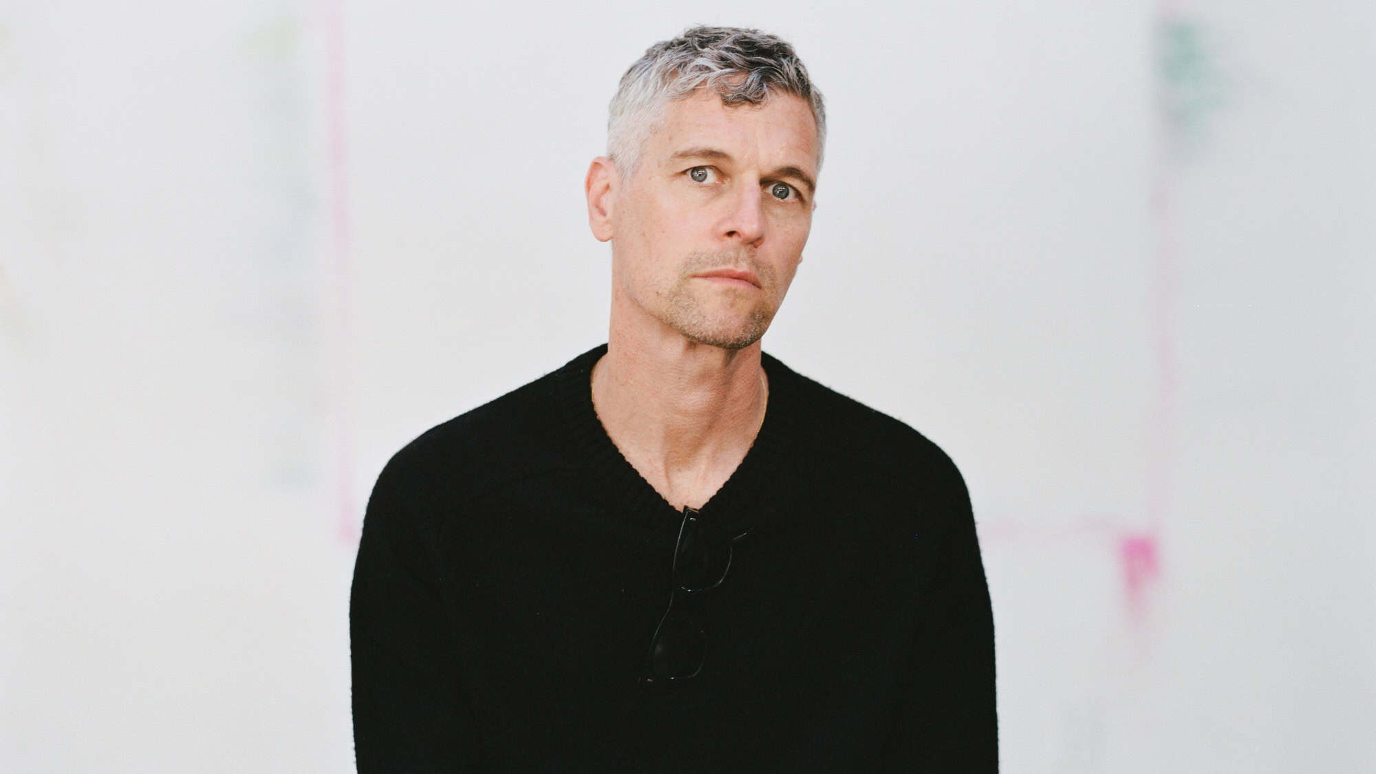 A man with short gray hair wearing a black sweater and glasses sits in front of a plain white background.