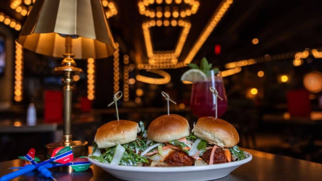 A plate of three chicken sliders with lettuce is on a table next to a cocktail and a gold lamp in a dimly lit restaurant with decorative lights in the background.