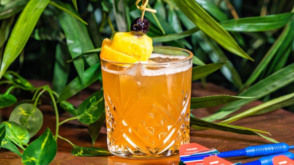 A glass of orange cocktail with ice, garnished with orange slices and a cherry, sits on a wooden surface with green leaves and darts nearby.