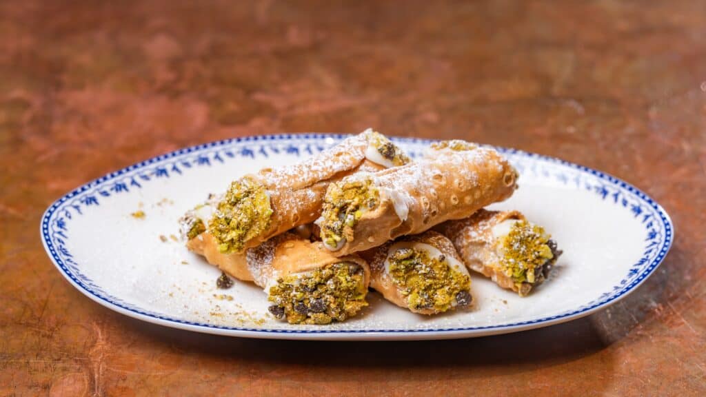 A plate of six cannoli filled with cream and topped with chopped pistachios, dusted with powdered sugar, on a white and blue patterned dish.