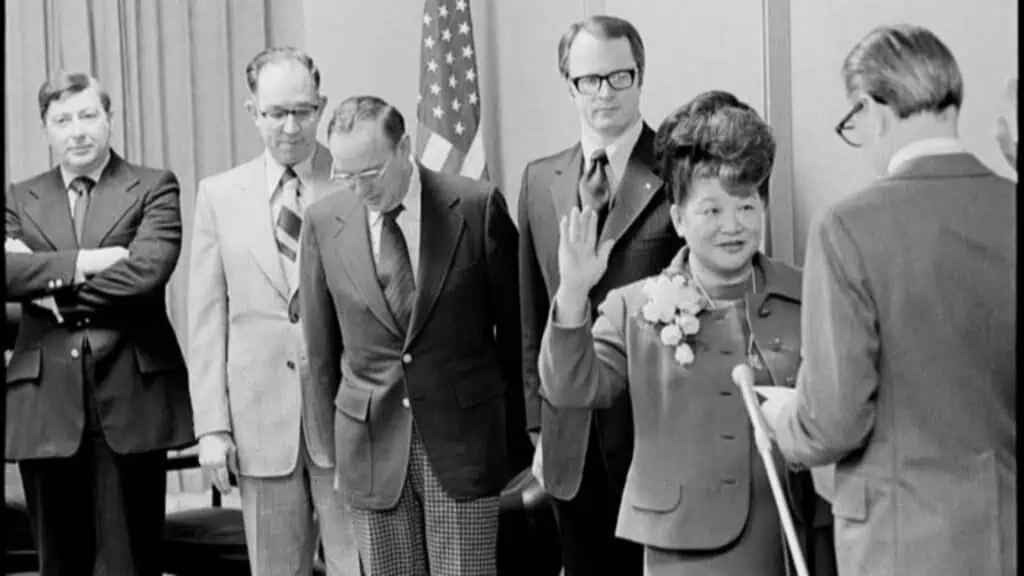 A woman with a corsage raises her right hand during a swearing-in ceremony in Seattle, making history as several men in suits stand nearby; an American flag is visible in the background.