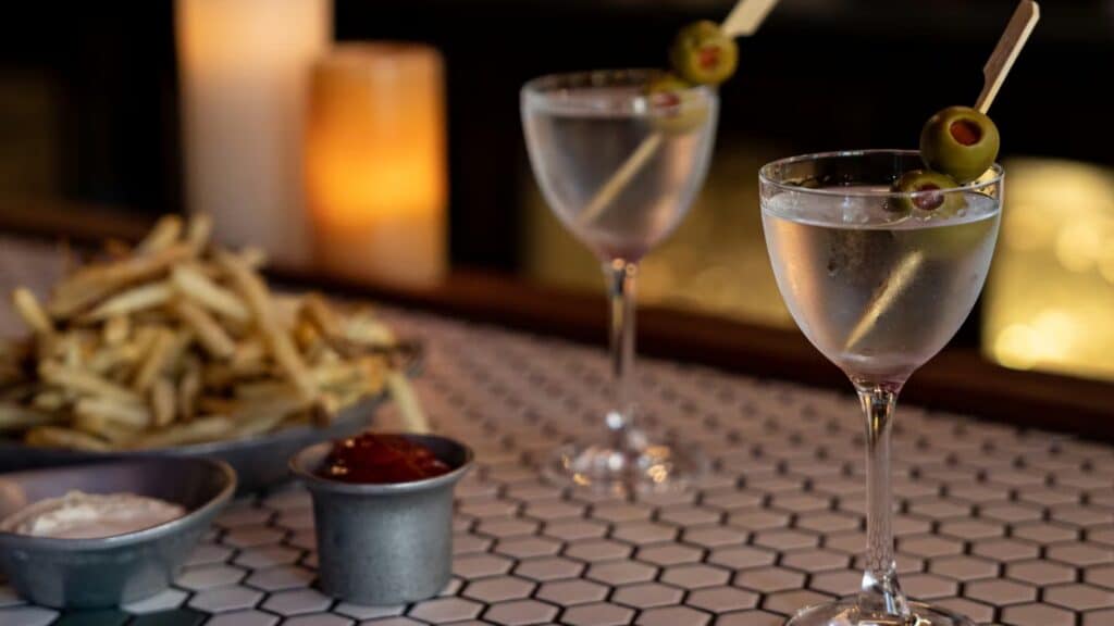 Two martini glasses garnished with olives sit on a hexagonal tile bar next to a plate of fries with ketchup and dipping sauce; candles are in the background.