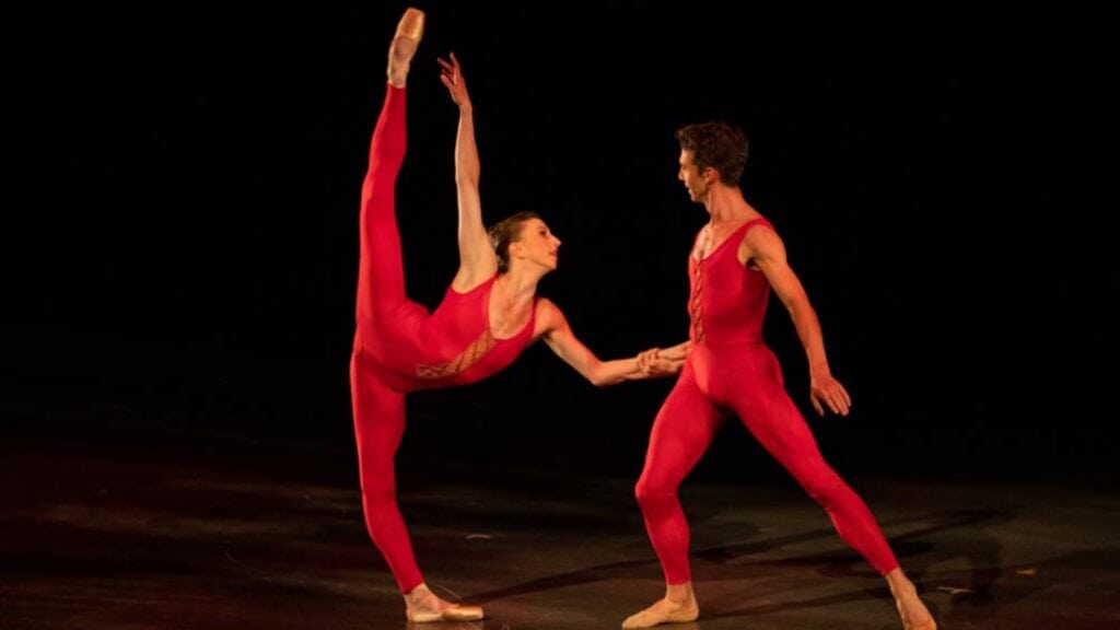 Two ballet dancers in matching red costumes perform on stage; one dancer lifts a leg high while holding the other's hand, bringing a touch of Arty Anniversary elegance to the performance.