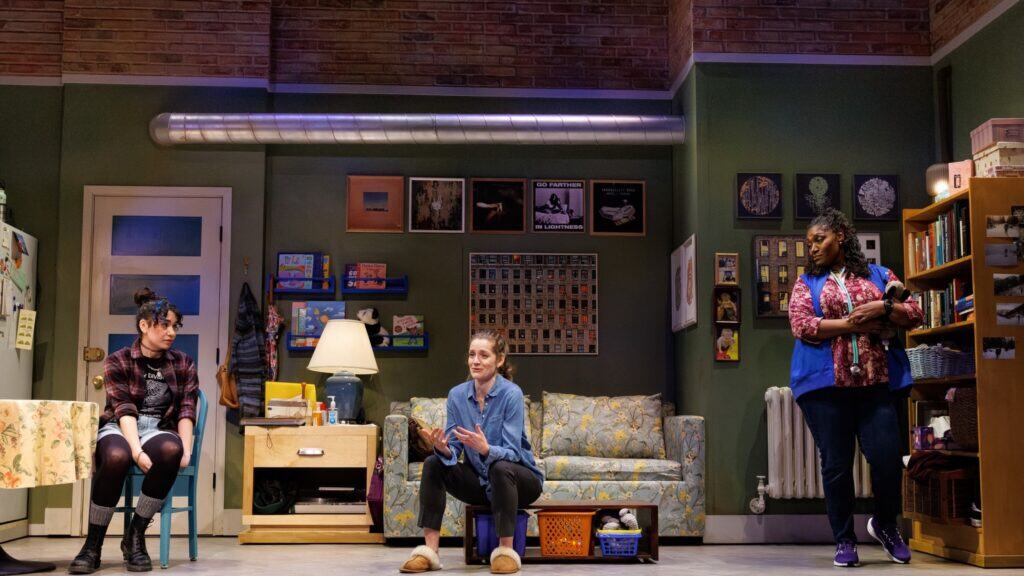 Three women are in a living room set on stage at Seattle Rep, with two seated and one standing near a bookshelf, surrounded by books, wall art, and household items—a scene that feels like looking in the rearview mirror.