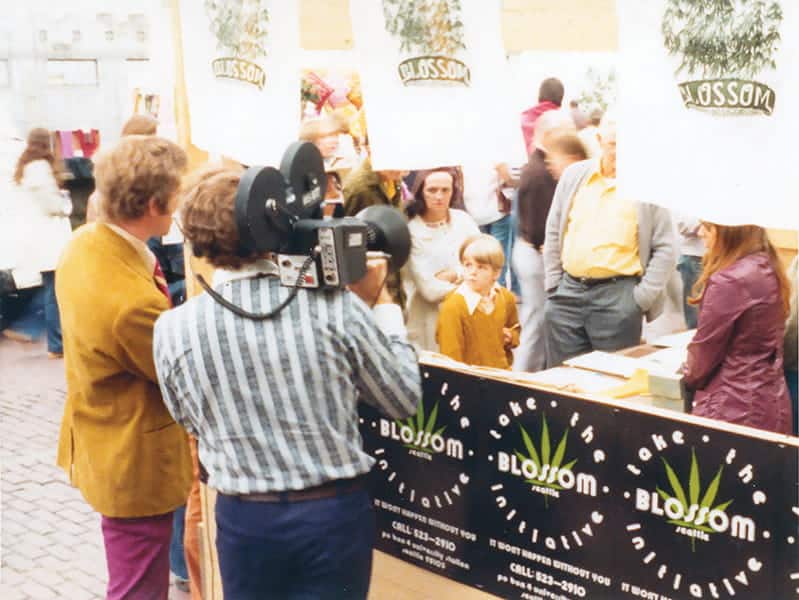 A cameraman films a boy and several adults at an indoor market stall decorated with “Blossom” banners, posters, and artifacts. People are standing and observing the scene as excitement rises Up In Smoke.