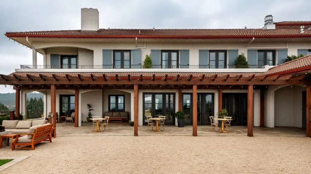 Two-story building with a wooden pergola, outdoor seating, and gravel patio area, set against a cloudy sky.