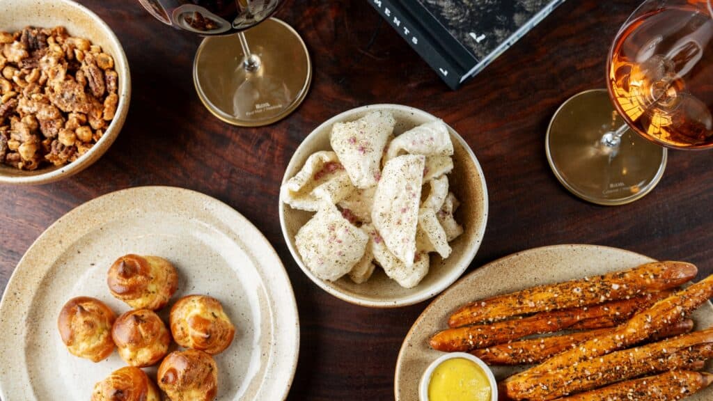 Bowls of snacks including seasoned nuts, puffed chips, pretzel sticks with mustard, and bite-sized rolls on a wooden table, accompanied by two glasses of wine.