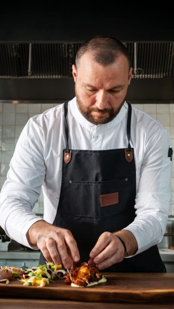 A chef in a white shirt and dark apron carefully plating food on a wooden board in a professional kitchen.