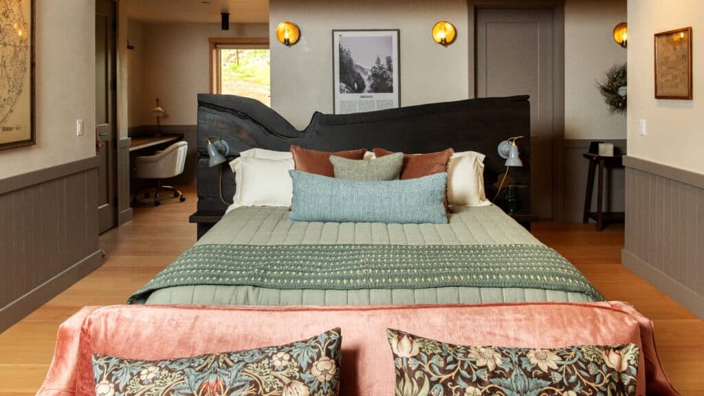A neatly made bed with green bedding and decorative pillows faces a pink sofa with patterned cushions in a modern bedroom with wood accents and wall art.