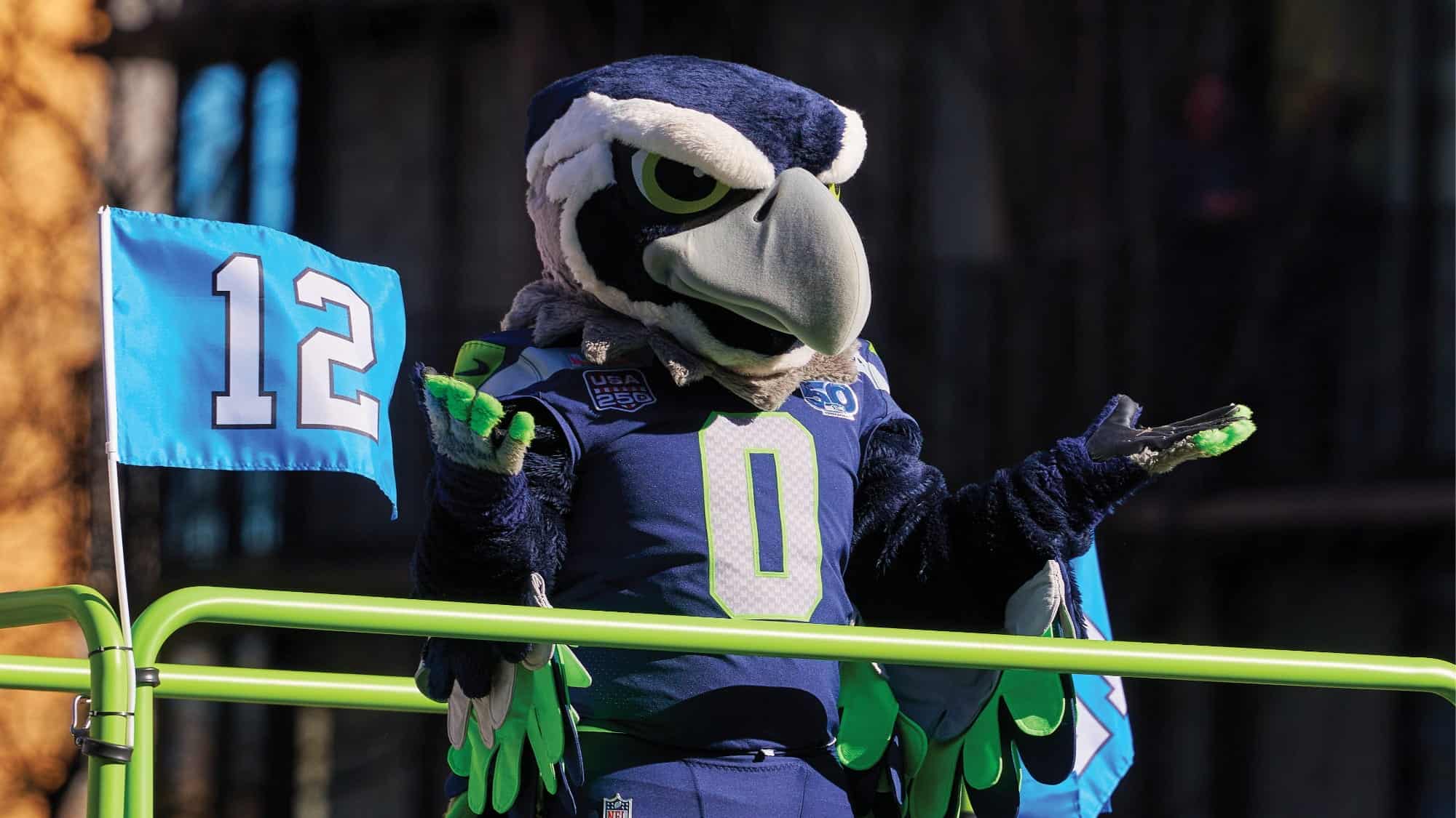 The Seattle Seahawks mascot, dressed in a blue and green costume, stands next to a blue NFL flag with the number 12, gesturing with both hands raised, celebrating the team's Super Bowl LX spirit.