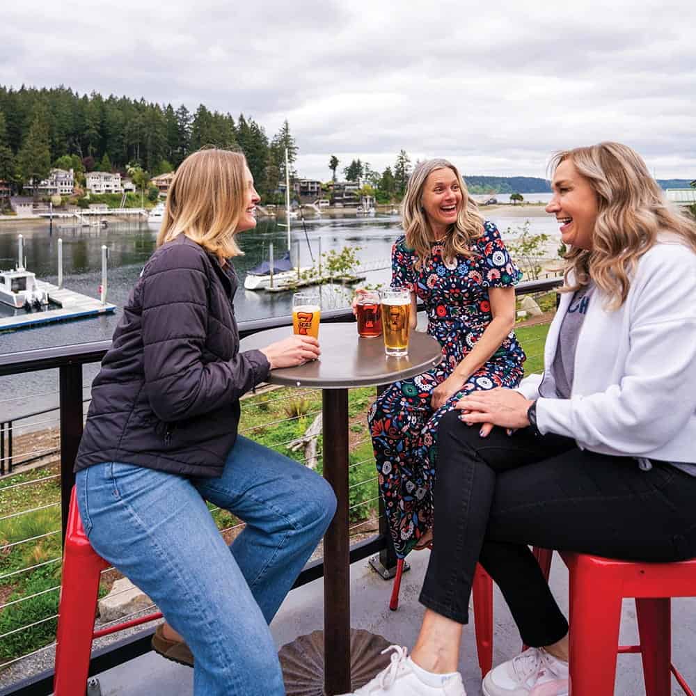 Three women sit around a high table with drinks, laughing together on an outdoor patio overlooking the marina and trees in Gig Harbor—one of the best places to live.