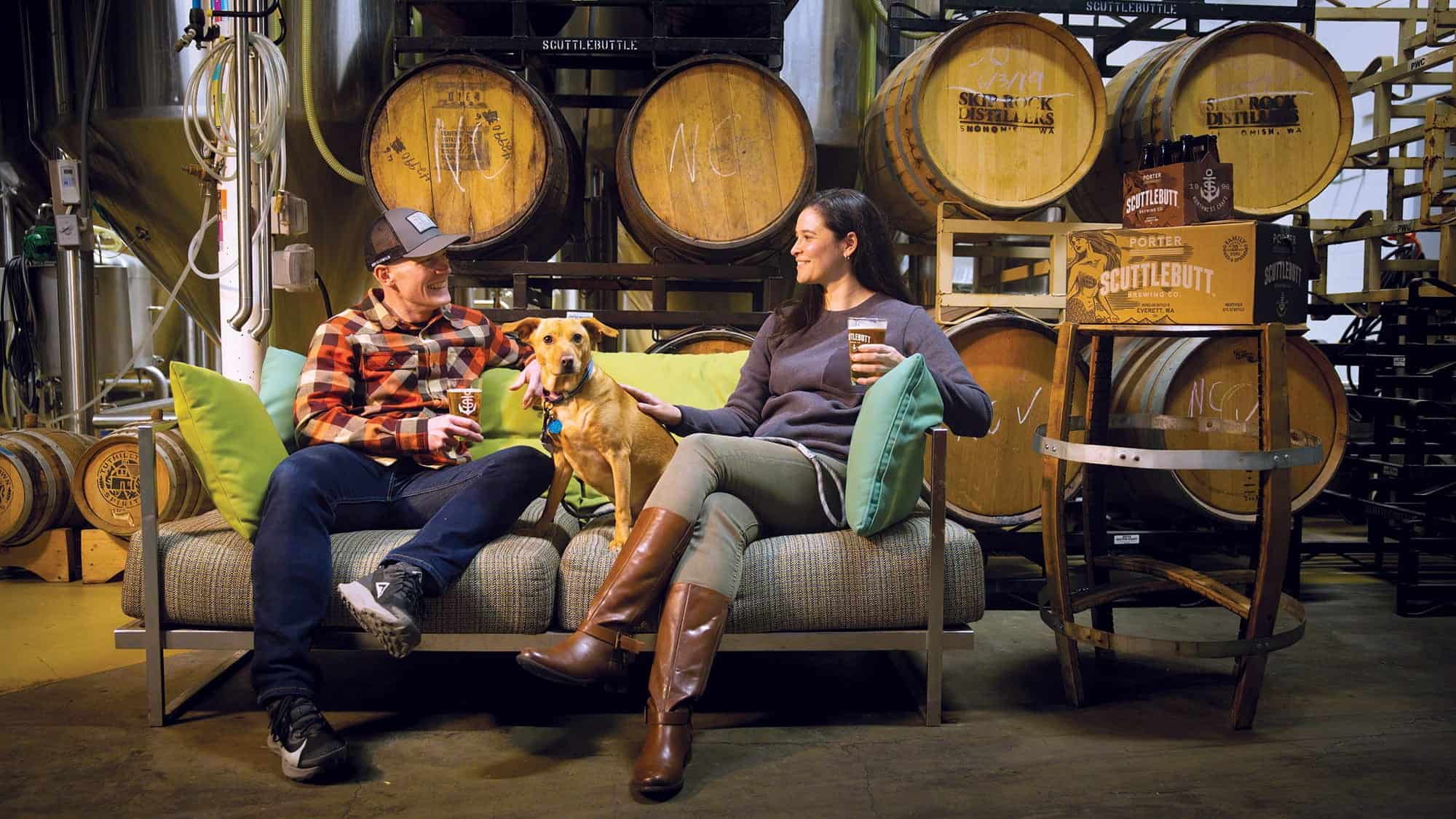 Two people sit on a couch with a dog, holding drinks, in a brewery with wooden barrels and boxes labeled "Scuttlebutt" in Everett—one of the Best Places to Live—creating a cozy, welcoming vibe.