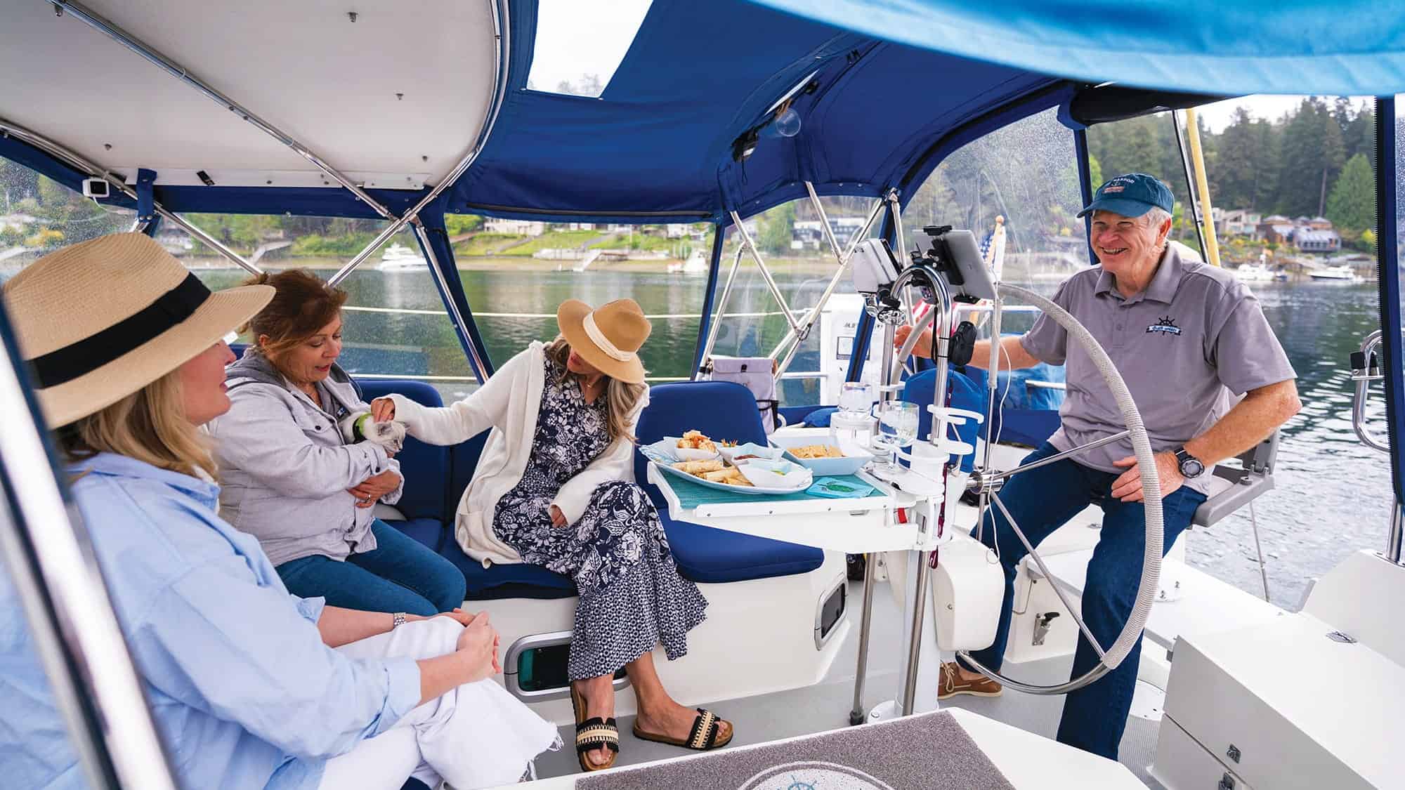 Four people sit and chat on a sailboat under a blue canopy, sharing food at a table with water and Gig Harbor homes in the background, capturing the relaxed atmosphere of one of the Best Places to Live.