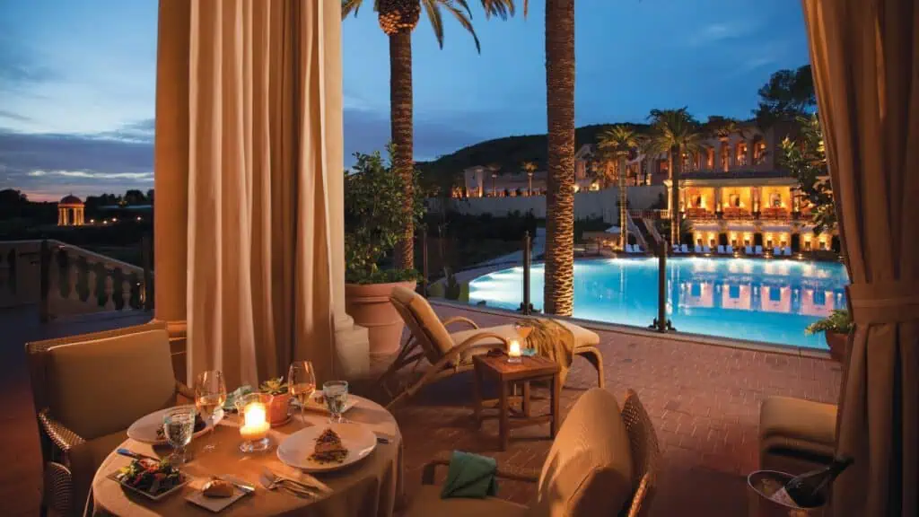 Table set for two with dinner and dessert beside a lit candle, overlooking a pool and illuminated building at dusk, framed by palm trees and curtains.