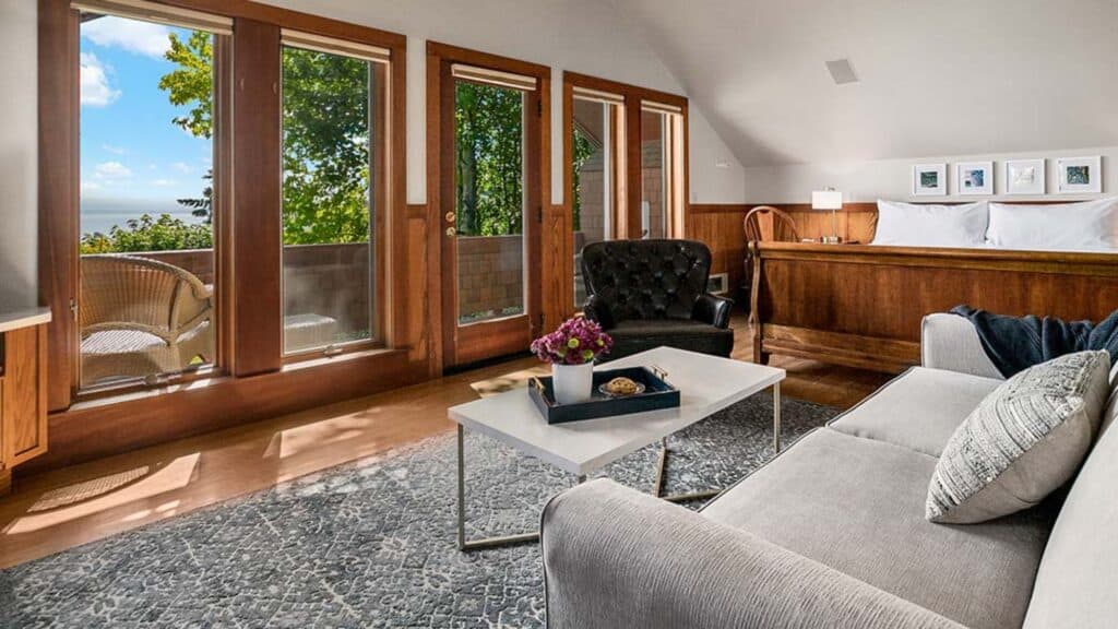 A bright, modern living area with a gray sofa, armchair, white coffee table, and large windows leading to a balcony with outdoor seating and a view of trees and water.