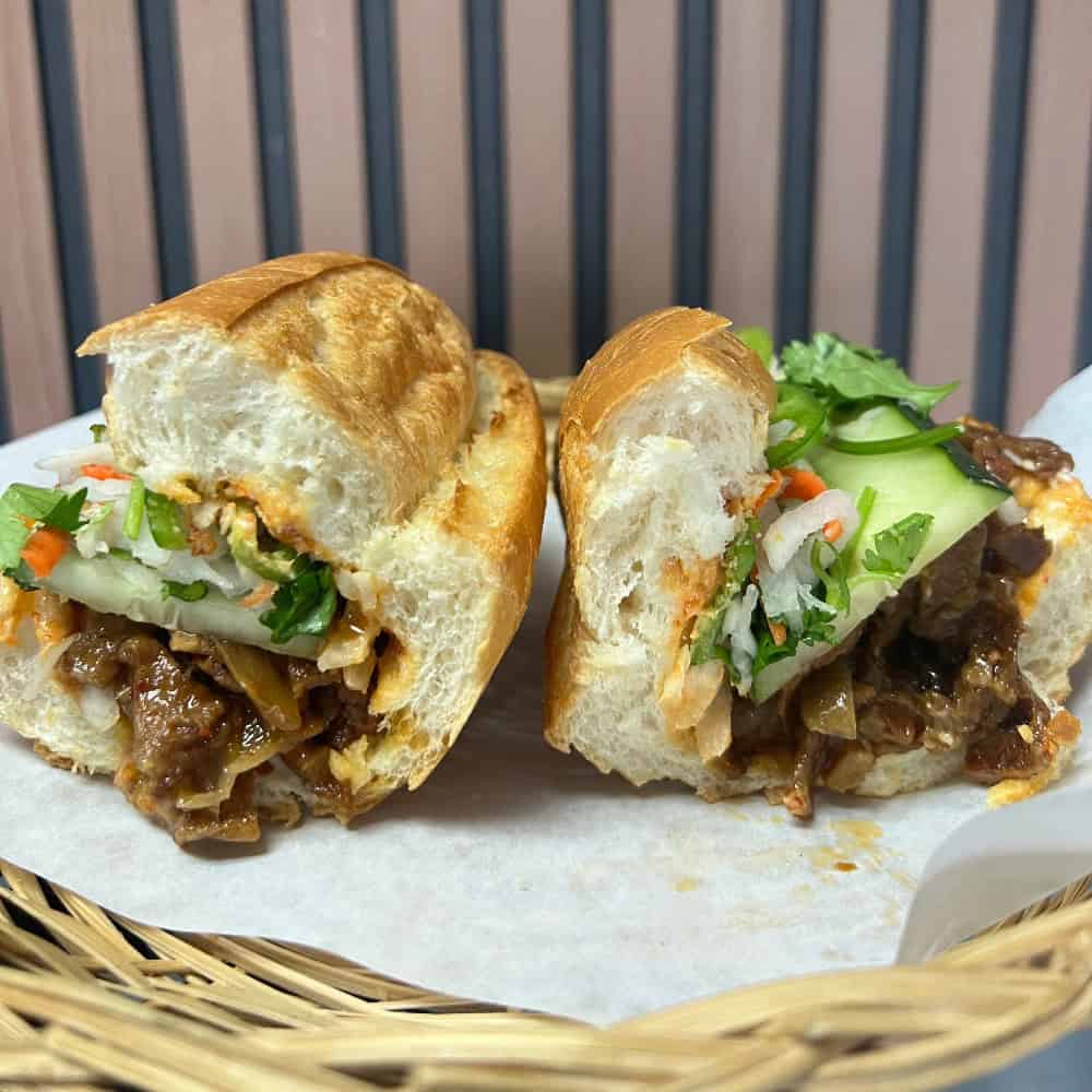 A sliced banh mi sandwich on a paper-lined basket, filled with meat, pickled vegetables, cucumber, and cilantro.