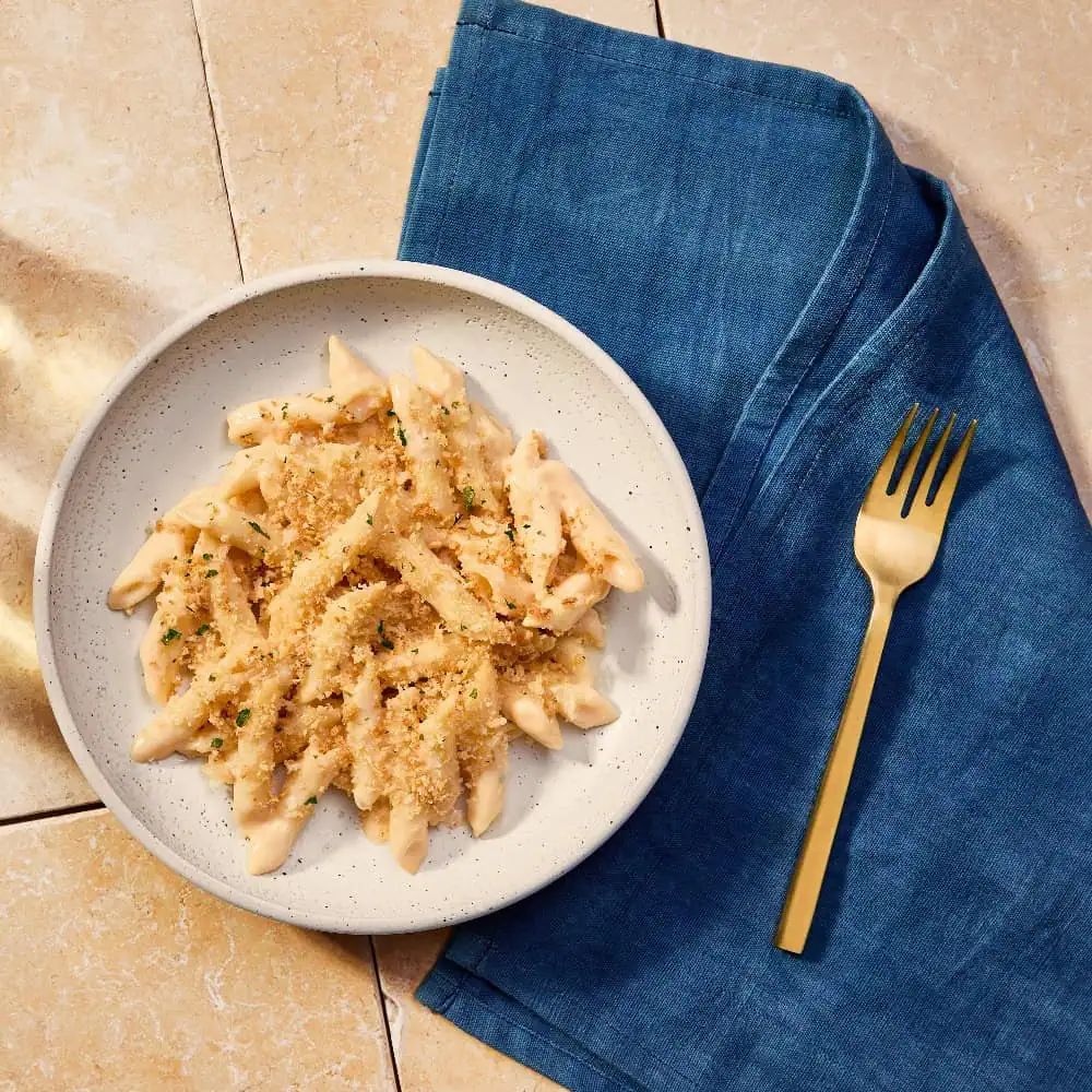 A plate of penne pasta with creamy sauce and breadcrumbs sits on a tiled surface next to a gold fork and a folded blue napkin.