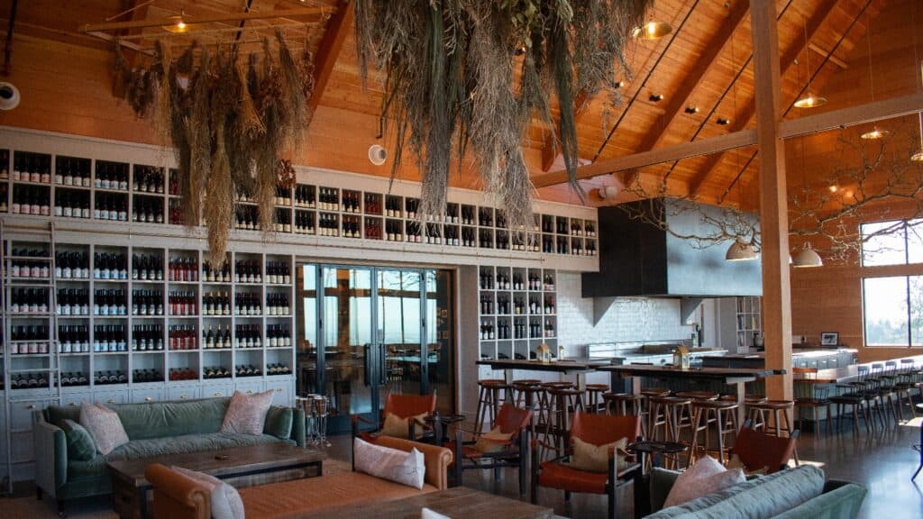 Modern winery tasting room with high wooden ceilings, hanging dried plants, shelves filled with wine bottles, cozy seating, and a bar with stools.
