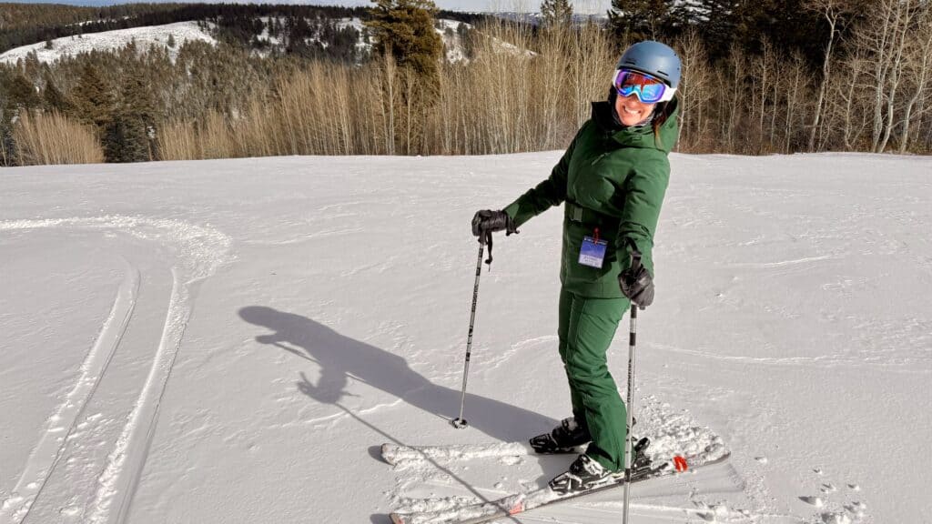 Person in green ski outfit and helmet standing on skis in the snow, holding ski poles, with trees and hills in the background.