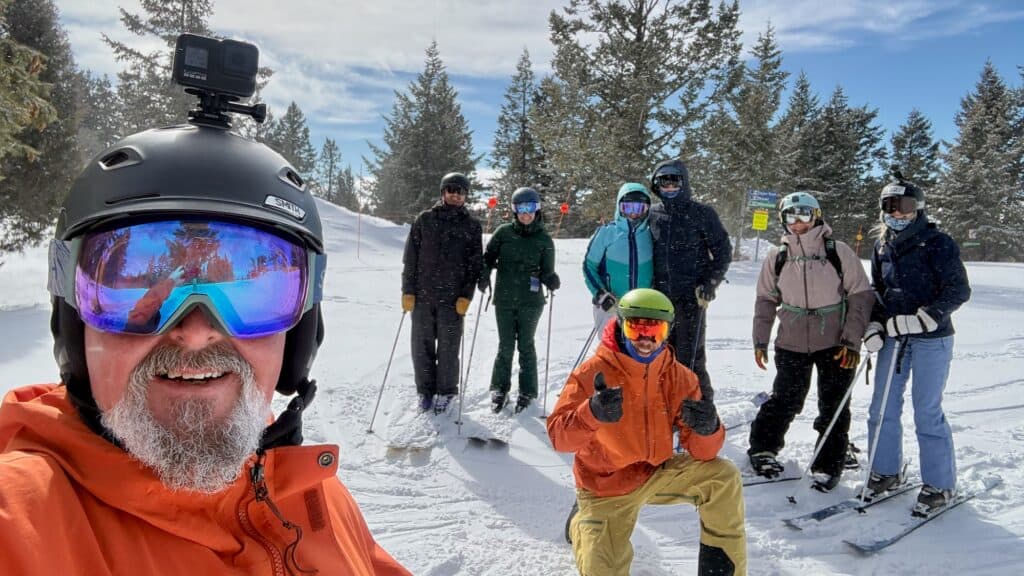 A group of seven skiers pose on a snowy slope, with trees in the background. One person in the foreground wears a helmet with a mounted camera and orange jacket.