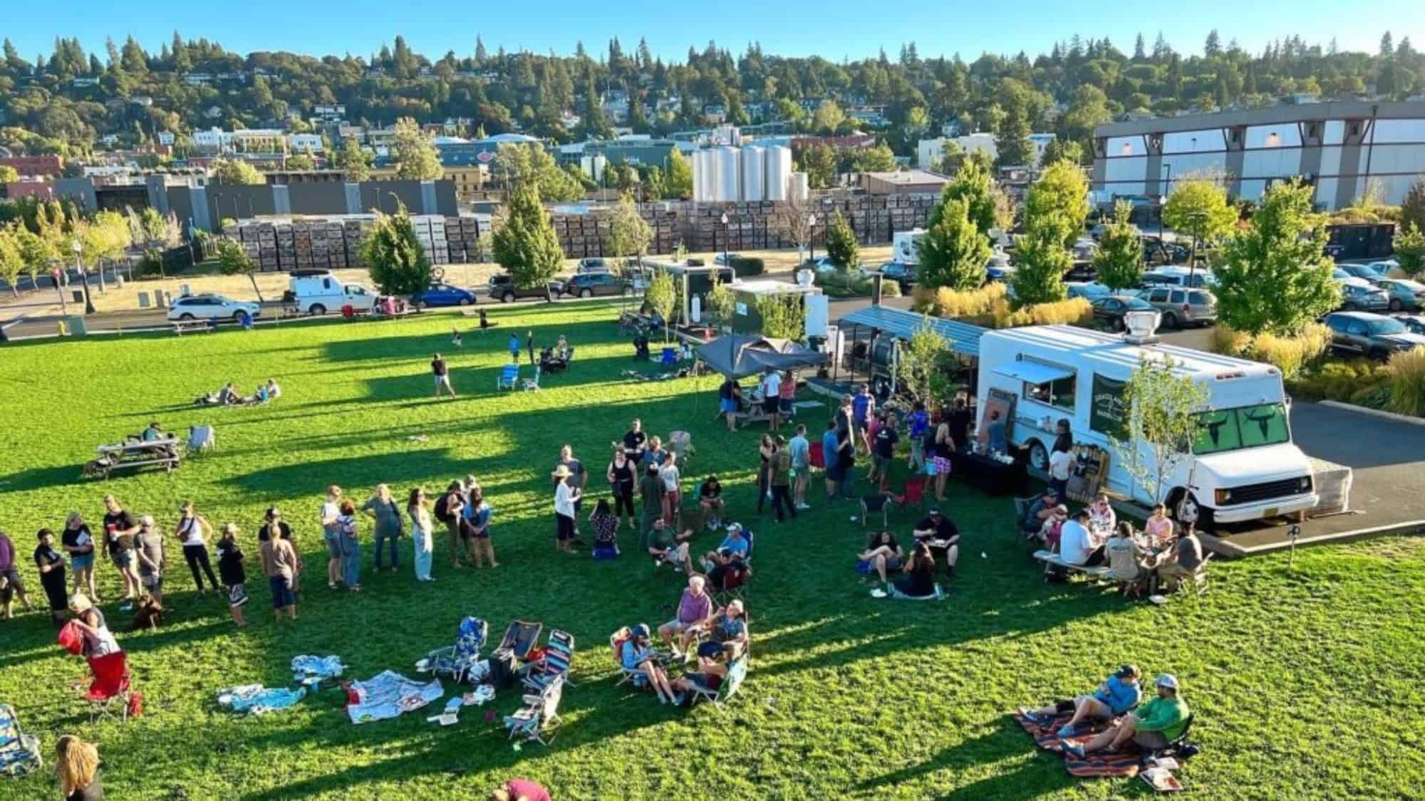 A crowd gathers on a grassy field near a Baseball Bites food truck, with groups sitting on blankets and enjoying smoked brisket. Trees, buildings, and hills form the backdrop under a clear sky.