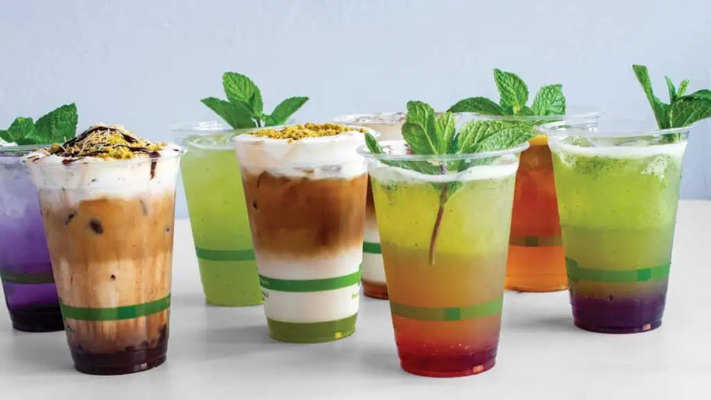 Assorted colorful iced drinks in clear plastic cups, some with cream, layered effects, and mint garnishes—perfect refreshments when exploring new spots to eat and drink in Seattle—displayed on a white surface against a light background.